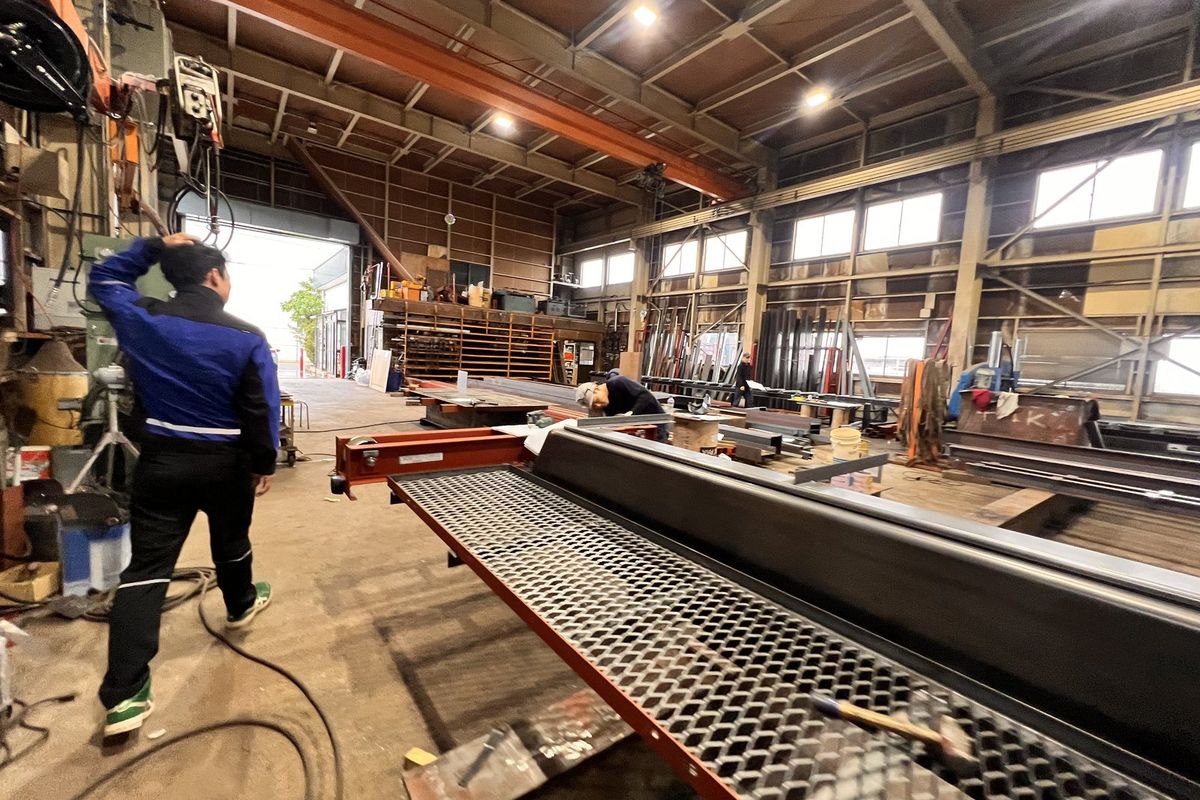 長い鉄骨の部材が置かれた工場の通路を歩く社員